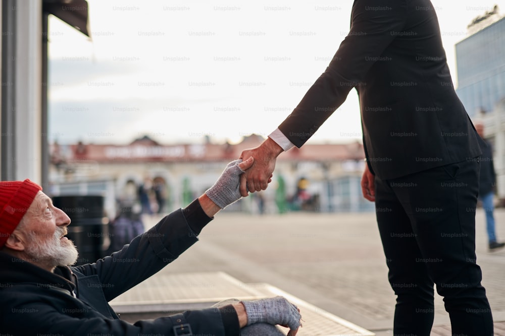 Mendigo mayor sentado al lado de la calle con solicitud de ayuda para recibir algunas monedas de un amable hombre de negocios. Hombre extiende la mano a las personas sin hogar.