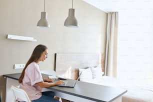 Pleasant pretty brunette girl wears light colourd t shirt and blue jeans, sitting alone in big bright bedroom, working at table using laptop computer, right side shot