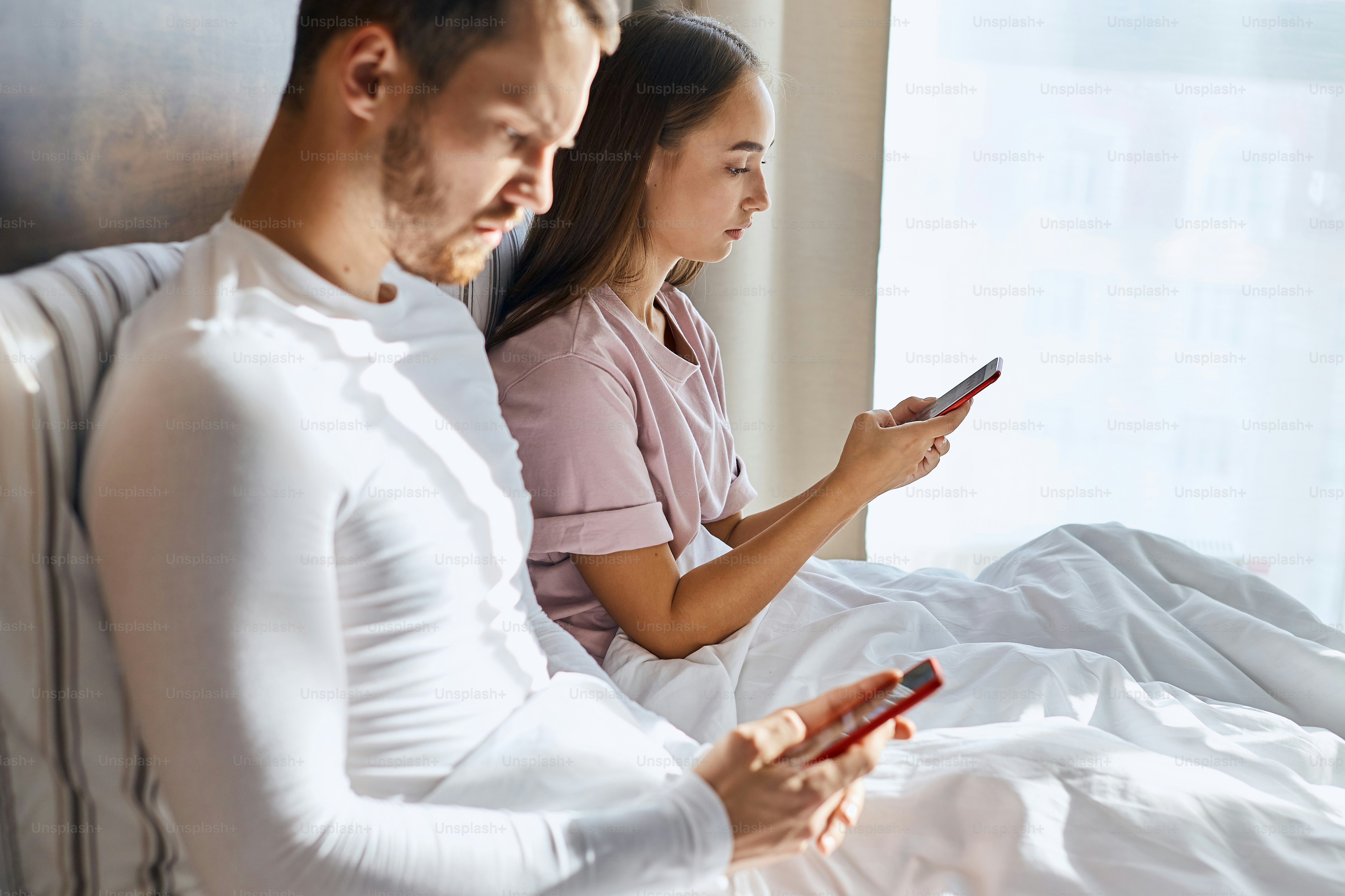 Close up of pretty attractive woman sitting on bed, covered with blanket, holding cell phone, chatting with interest, staring at screen, ignore live communication, family portrait