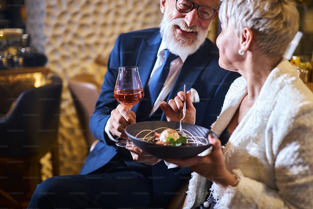 Kaukasischer Mann in Anzug und Brille, schöne Frau in weißem elegantem Blazer genießen Essen an einem attraktiven Ort. Süßes Dessert und ein Glas Champagner. Liebe liegt in der Luft