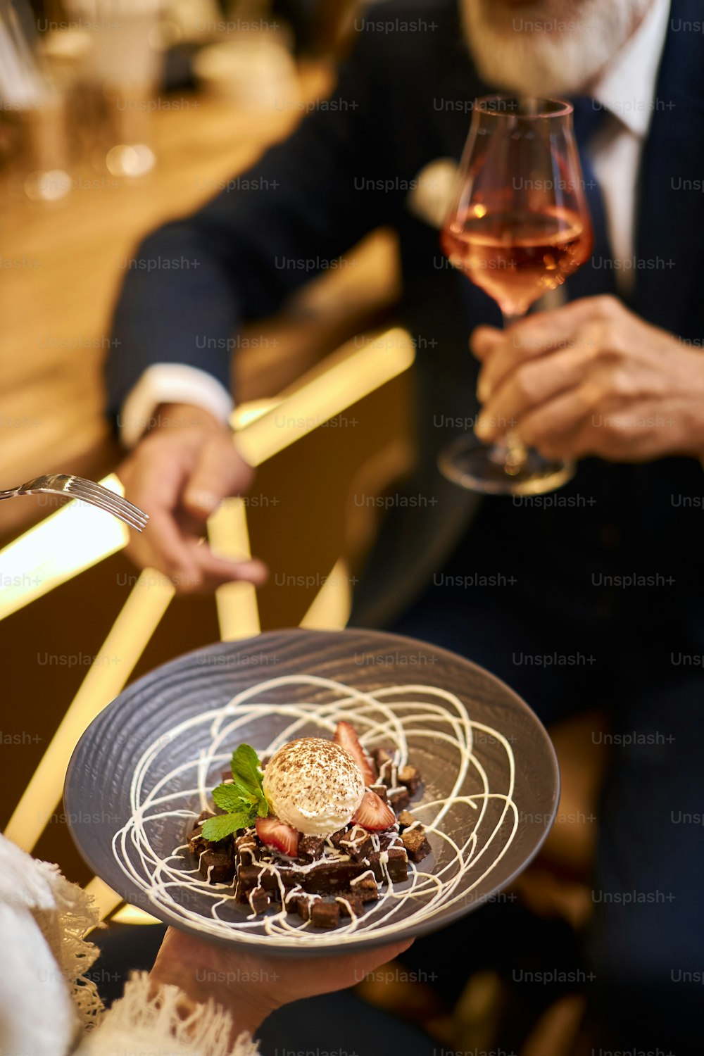 Imagem próxima da mão com taça de champanhe, sobremesa doce no prato cinza. Homem barbudo em smoking bebendo
