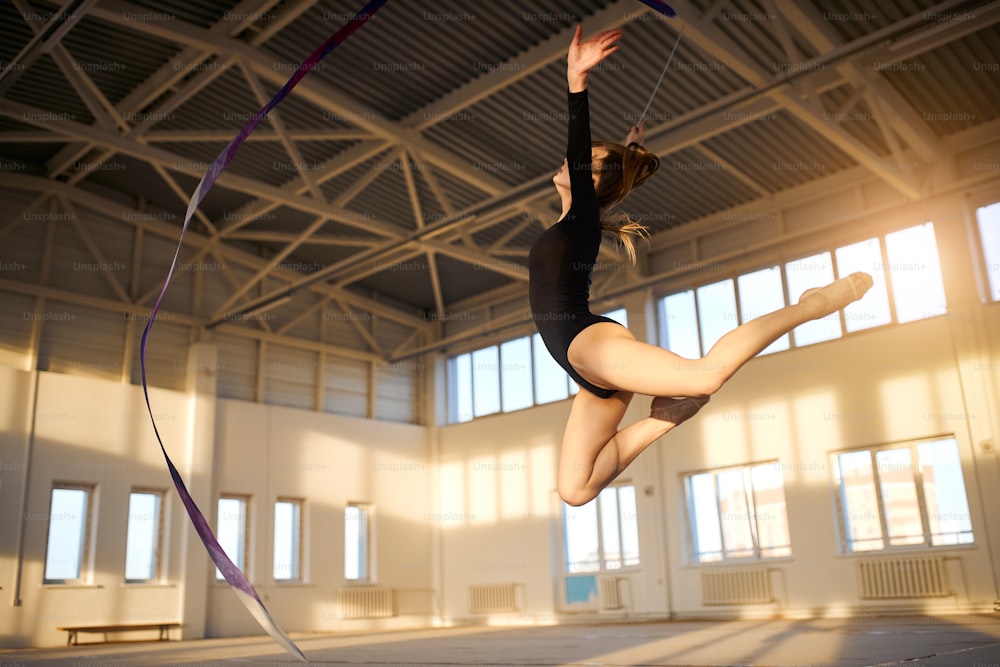 Gracieuse fille sportive sautant haut avec un long ruban coloré dans les mains, les jambes étirées en l’air, concept de gymnastique artistique, prise d’en bas