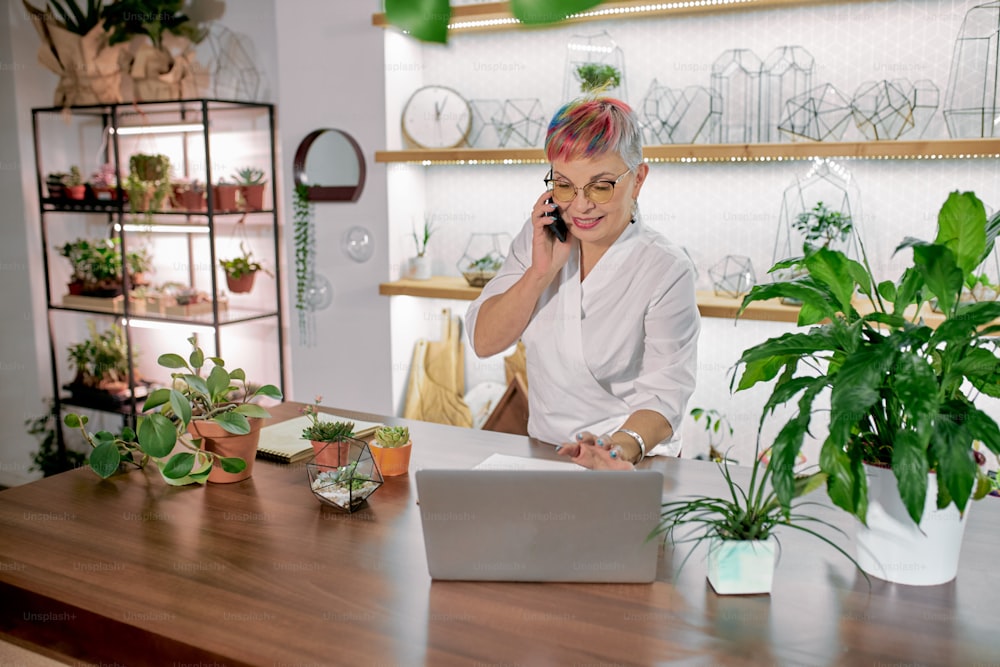 Modern short-haired senior woman engaged in floristy. Caucasian woman in white bathrobe stand talking on phone and using laptop, light room background