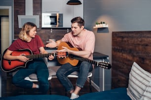 Two caucasian young people wearing casual clothes sit friendly talking, have conversation holding guitars at home