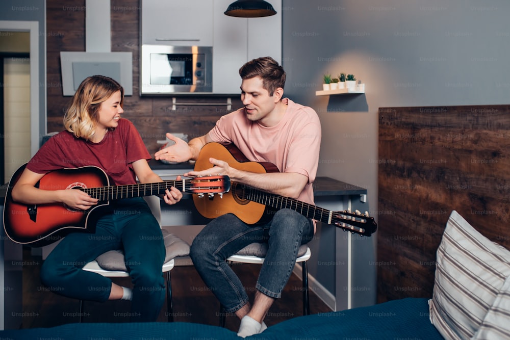 Two caucasian young people wearing casual clothes sit friendly talking, have conversation holding guitars at home