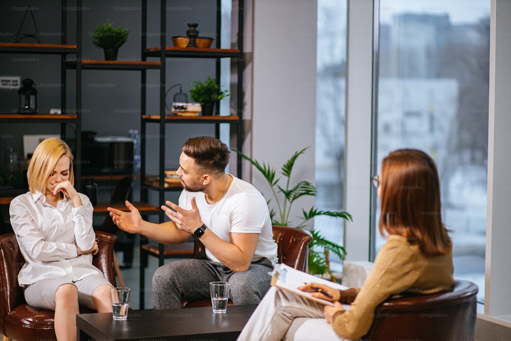 family disagreement, people relationships concept. young caucasian married couple discussing their problems at marital therapy with psychologist, psychologist sit attentively listen to irritated couple and give advice, support, possible solutions to their problems.