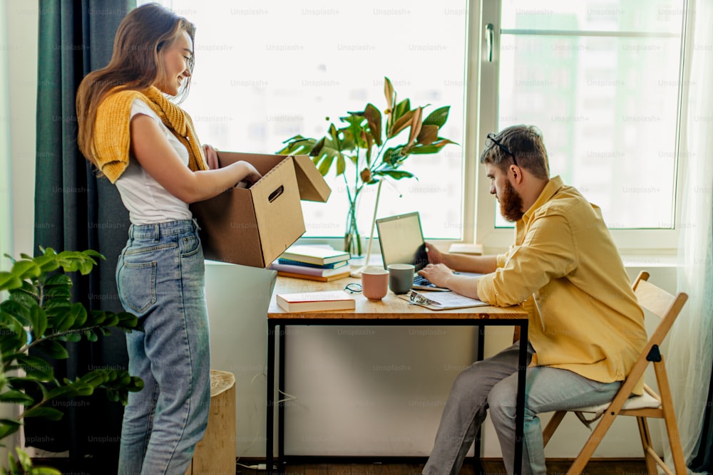 La mujer se para junto a la mesa con la caja, hace las tareas domésticas, desempaca las cajas después de mudarse a una nueva casa mientras su esposo trabaja con la computadora portátil en casa, el hombre en el freelance. Pareja casada en apartamento nuevo