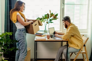 woman stand next to table with box, do household chores, unpacks boxes after moving into new house while her husband work with laptop at home, man at freelance. married couple in new apartment