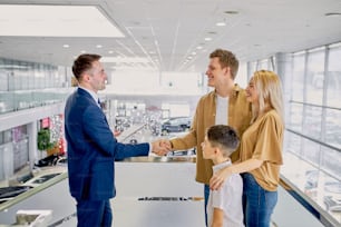 Agréable consultant caucasien heureux d’aider les clients dans la salle d’exposition de voitures, beau vendeur en costume formel avoir une conversation avec les clients