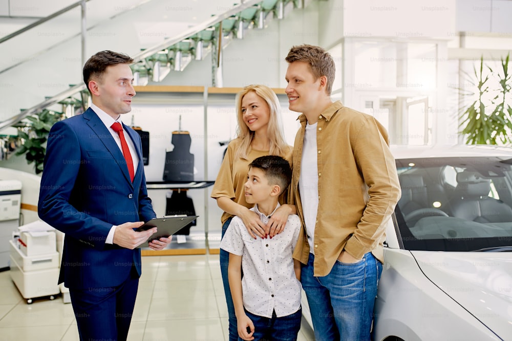 affable caucasian salesman consult customers in cars dealership. young family came to make purchase, to get new car, they listen to young consultant, he is talking about characteristics of auto