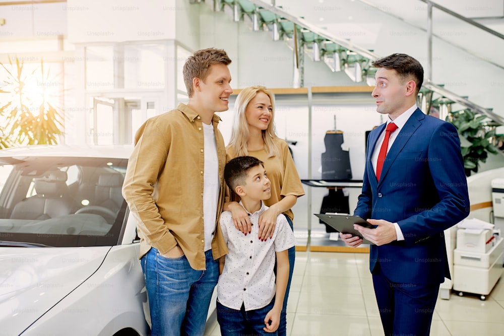 young beautiful caucasian couple with child choosing their first auto in dealership, customers look at variety of cars, try to choose the best, thinking. consultant help them, give consultation