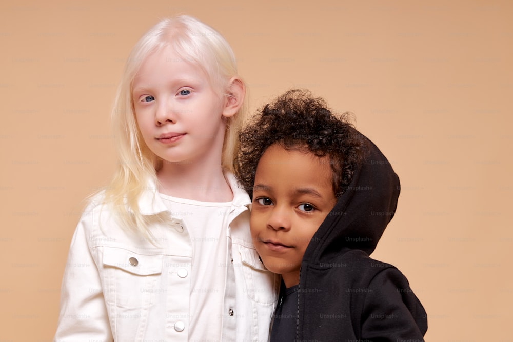 Niedliche entzückende verschiedene Kinder, die vor der Kamera posieren, kleines ungewöhnliches Albino-Mädchen mit dunkelhäutigem multirassischem Jungen, der auf beigem Hintergrund isoliert ist