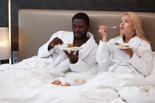 귀여운 커플은 침대에서 함께 아침을 먹고, 사랑스러운 백인 아가씨는 아프리카 남편과 행복해하며, 그들은 사랑에 빠졌다