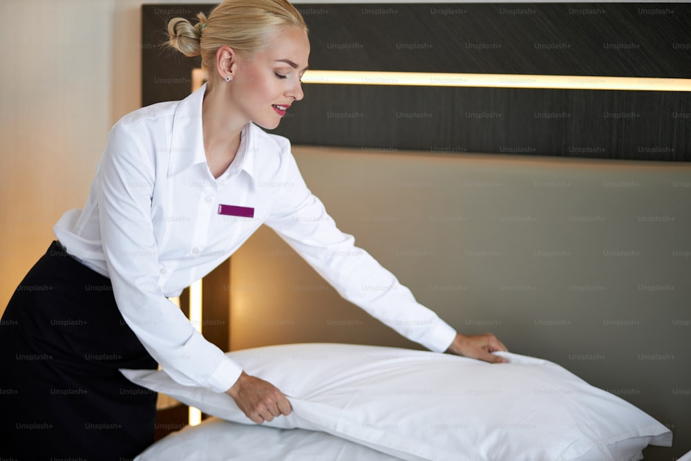 beautiful young hotel maid setting up white pillow on bed sheet in hotel room, smile, enjoy