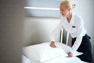 Hermosa criada cambia la funda de almohada en la cama, mujer rubia caucásica en uniforme prepara habitación para los huéspedes
