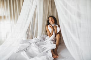 Beautiful tanned young woman awakening in white bed. Happy wake up and start new day. Leisure and rest. Wellbeing and carefree  concept. Long curly hair. Stay home.