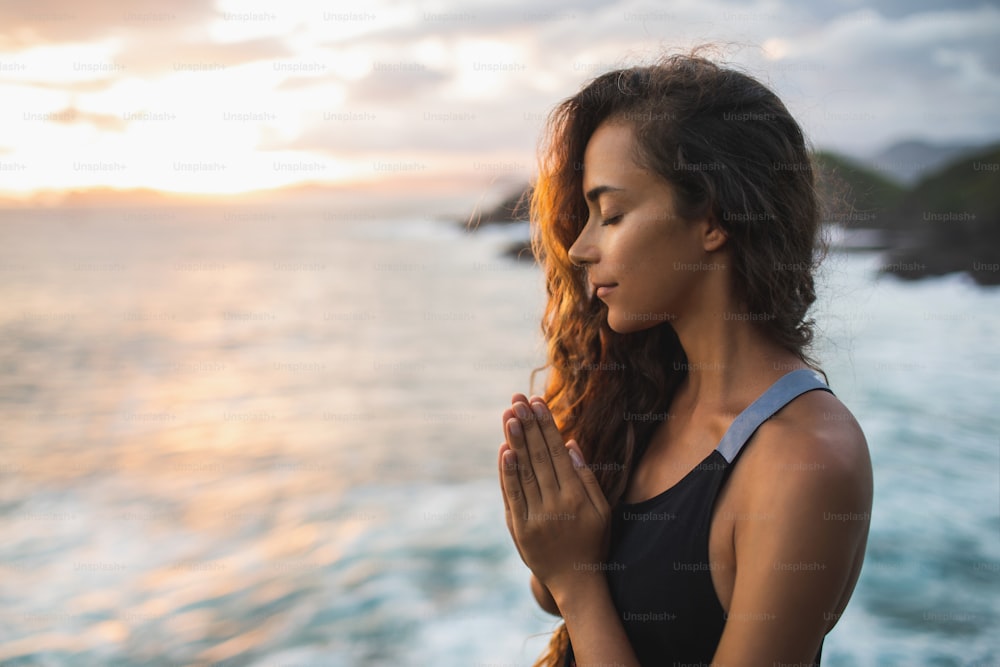 Young woman praying and meditating alone at sunset with beautiful ocean and mountain view. Self-analysis and soul-searching. Spiritual and emotional concept. Introspection, introversion and soul healing.