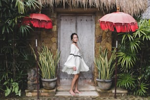 전통적인 발리 건축이 있는 우붓 마을의 하얀 튜닉을 입은 젊은 여성. 발리 하우스의 스타일입니다. 패션 스타일, 곱슬 머리, 가벼운 드레스. Changgu의 저택