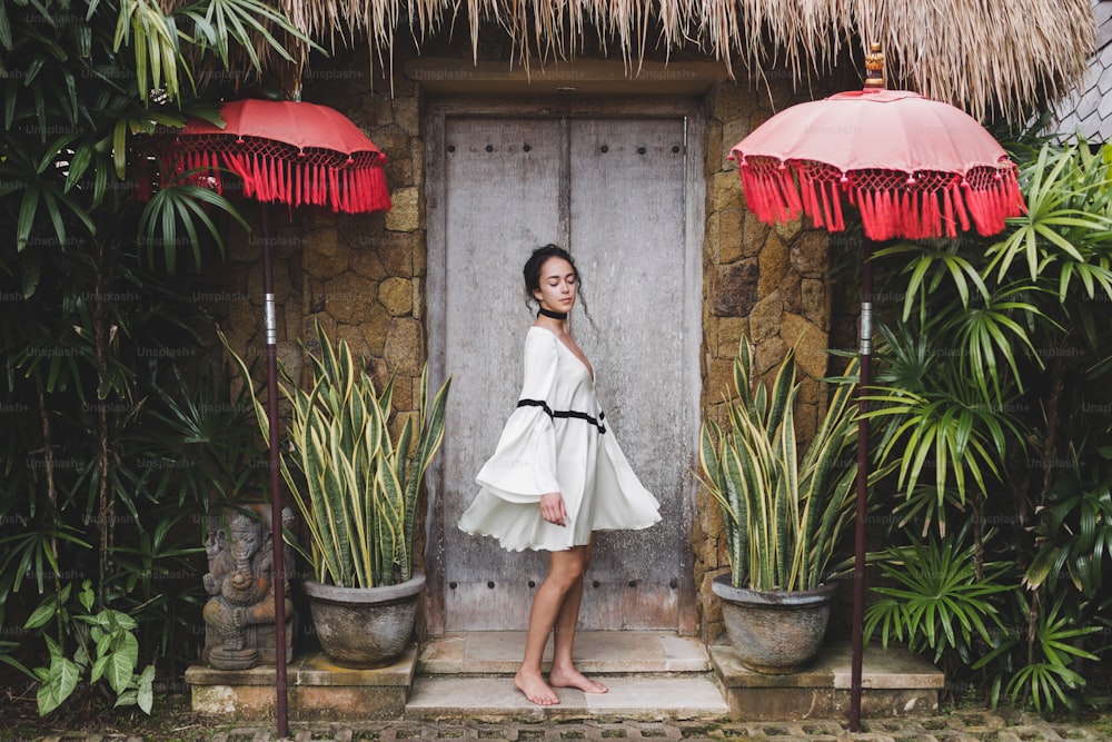 Young woman in white tunic in Ubud village with traditional balinese architecture. Style of Bali house. Fashion style, curly hair, light dress. Villa in Changgu