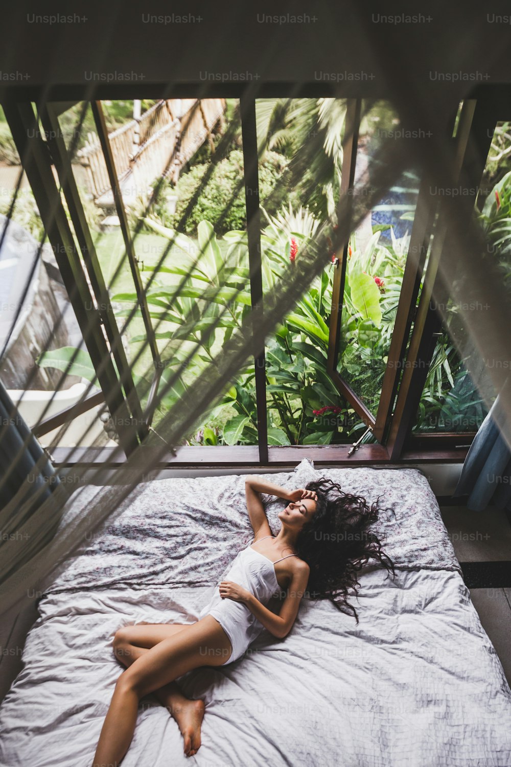 朝ベッドで目を覚ます女性、熱帯庭園の窓からの景色。ライフスタイル写真