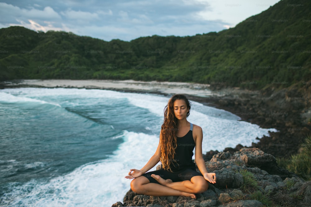 Mulher nova praticando yoga em pose de lótus ao pôr do sol com bela vista para o oceano e montanha. Sensibilidade à natureza. Autoanálise e busca da alma. Conceito espiritual e emocional.
