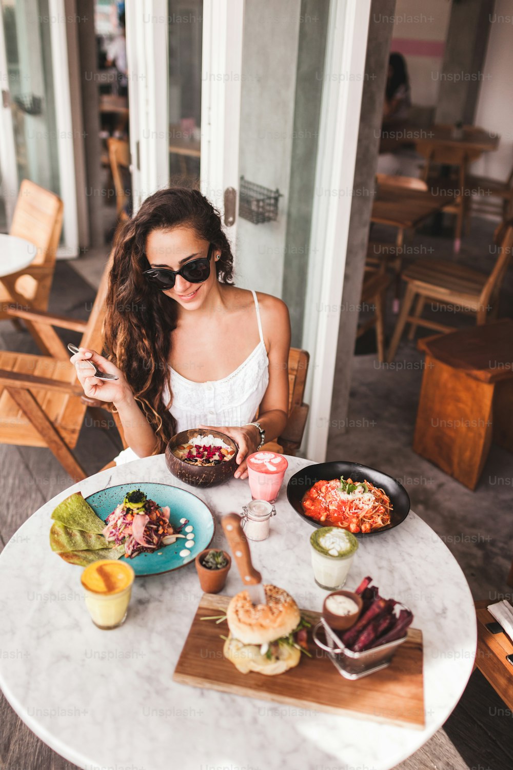 Young smiling woman having breakfast in stylish street cafe. Smoothie bowl, matcha latte, tacos and burger