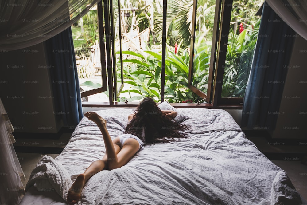 Donna che si sveglia nel letto al mattino, vista dalla finestra sul giardino tropicale. Foto di stile di vita