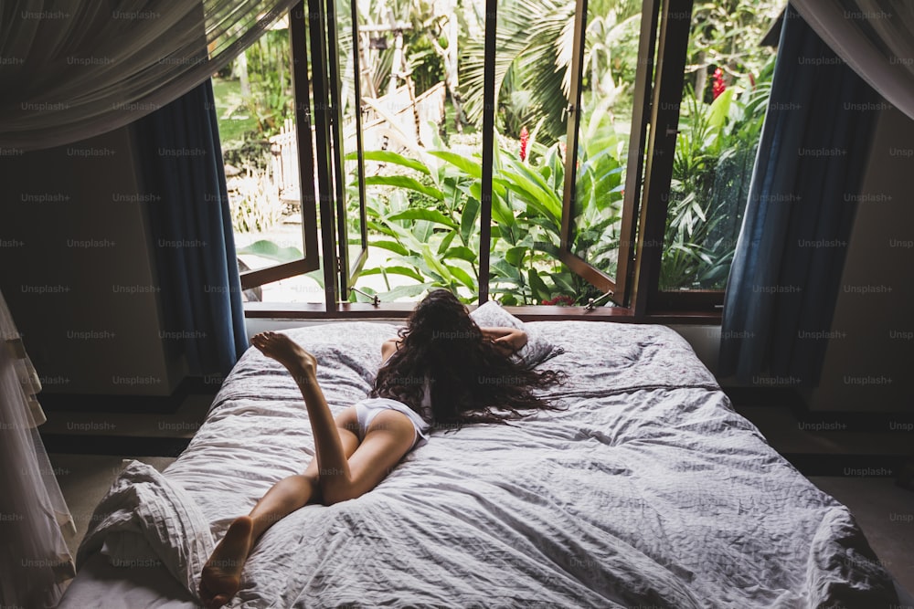 Frau, die morgens im Bett aufwacht, Blick aus dem Fenster auf den tropischen Garten. Lifestyle-Foto
