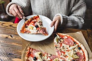 Le mani della donna mettono su un piatto un pezzo di pizza calda fresca con prosciutto, carciofi, olive e pomodori