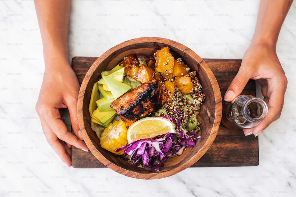 Woman eating meal with fried salmon fish steak, quinoa, avocado, corn, cabbage salad and baked pumpkin in wooden bowl. Healthy organic food concept. White marble table surface.