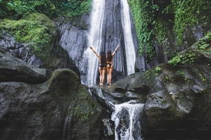 Two slim women relaxing near beautiful waterfall in Bali jungle. Nature adventure Dusun Kuning in Ubud area