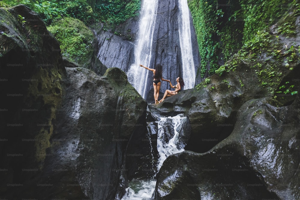 Two slim women relaxing near beautiful waterfall in Bali jungle. Nature adventure Dusun Kuning in Ubud area