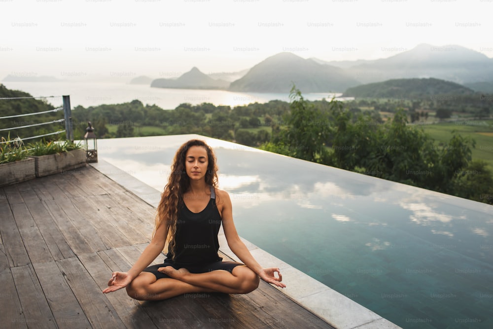Woman practicing yoga in lotus position on infinity poolside with beautiful ocean and mountain view in morning. Healthy lifestyle, spiritual and emotional concept. Awakening.
