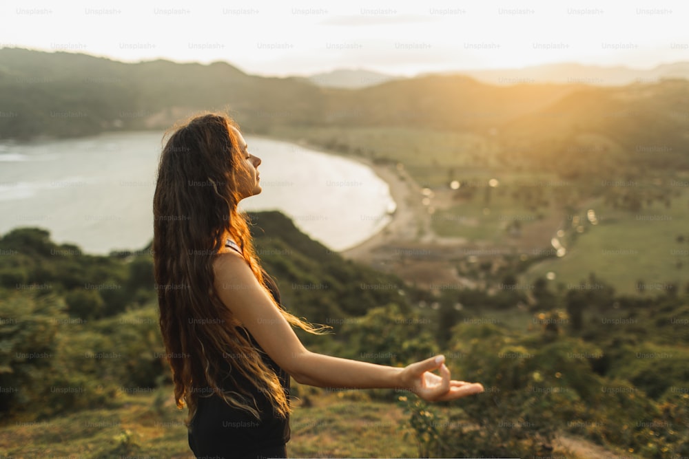 Giovane donna che pratica posizioni yoga al tramonto con splendida vista sull'oceano e sulle montagne. Sensibilità alla natura. Autoanalisi e esame di coscienza. Concetto spirituale ed emotivo.