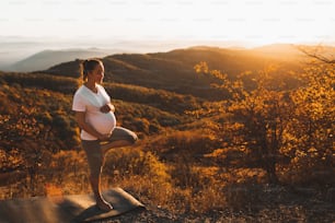 Concepto espiritual y emocional de armonía con la naturaleza en la época de maternidad. Mujer embarazada practicando yoga al aire libre en la colina al atardecer. Increíble vista otoñal a la montaña.