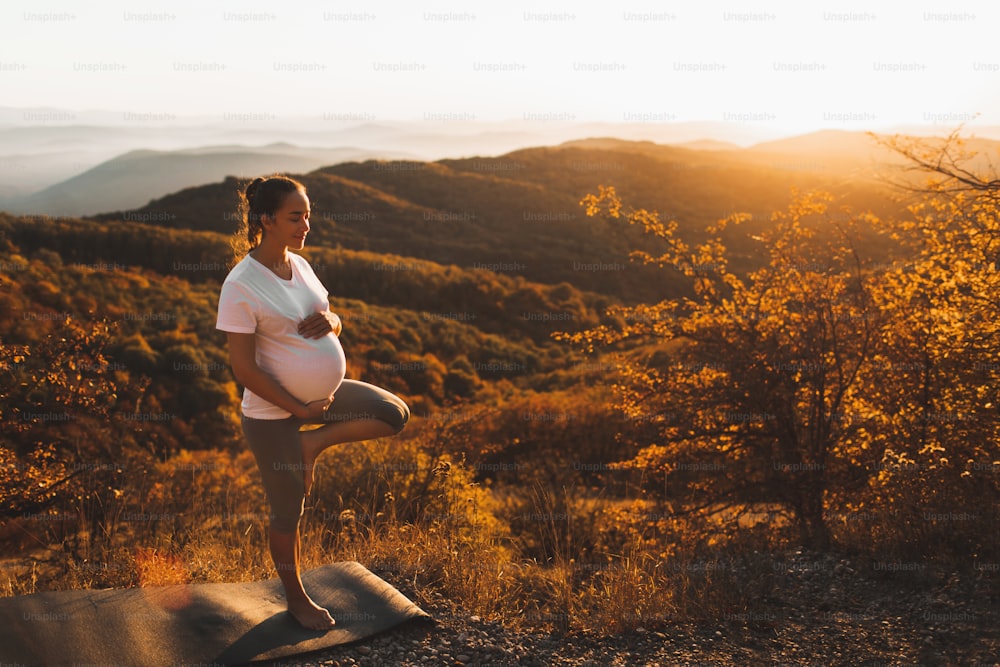 Conceito espiritual e emocional de harmonia com a natureza em tempo de maternidade. Mulher grávida praticando yoga ao ar livre na colina ao pôr do sol. Incrível vista para a montanha de outono.
