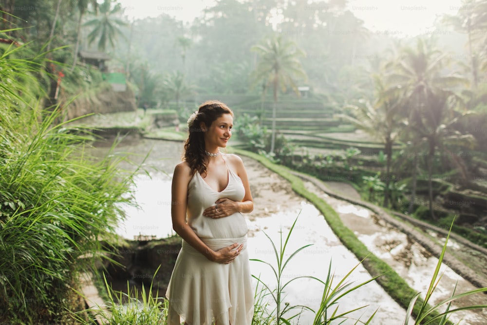 Young pregnant woman in white dress with view of Bali rice terraces in morning sunlight. Harmony with nature. Pregnancy concept.