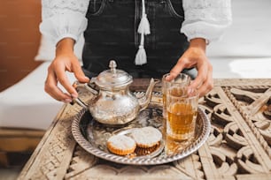 伝統的なモロッコのミントティーセレモニーをクッキーとヴィンテージの銀のティーポットで提供する女性の手。モロッコ、マラケシュのホスピタリティとサービス。