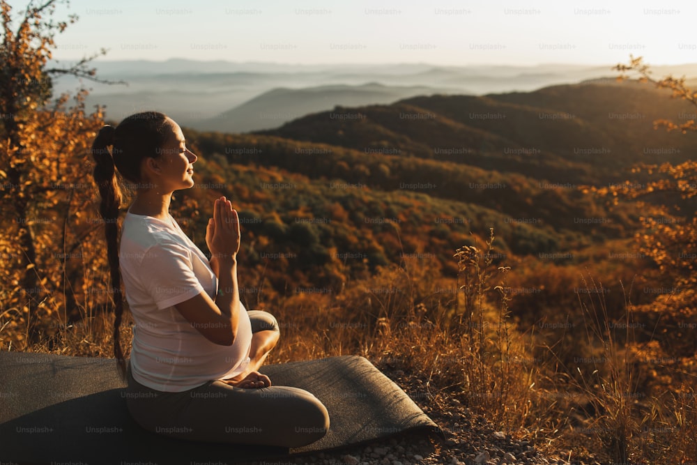 Conceito espiritual e emocional de harmonia com a natureza em tempo de maternidade. Mulher grávida orando sozinha ao ar livre na colina ao pôr do sol. Incrível vista para a montanha de outono.