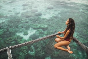 Bella donna abbronzata in costume da bagno rosso che si rilassa sull'amaca over-reef con splendida vista sulla barriera corallina in acqua trasparente. Vacanze in Asia concetto, sfondo tropicale.