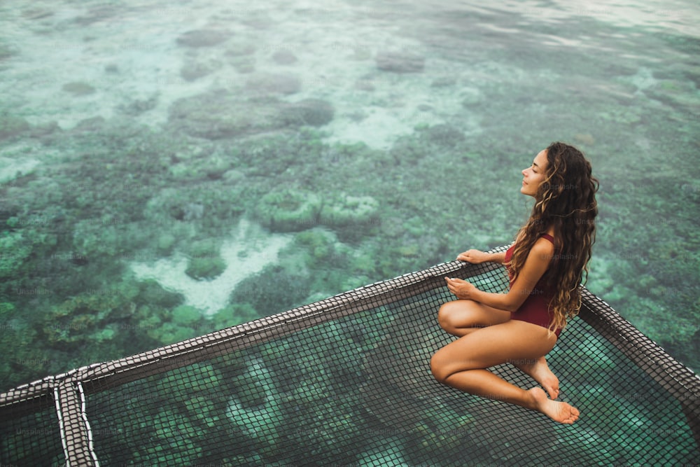 Beautiful tanned woman in red swimsuit relaxing in over-reef hammock with amazing view of coral reef in transparent water. Vacations in Asia concept, tropical background.