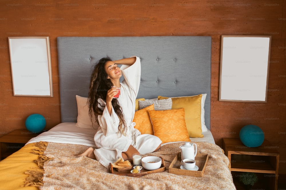 Mulher feliz em roupão branco desfrutando de café da manhã saudável na cama com granola, torradas crocantes, manteiga, leite e suco de melancia. Lazer e férias em hotel.