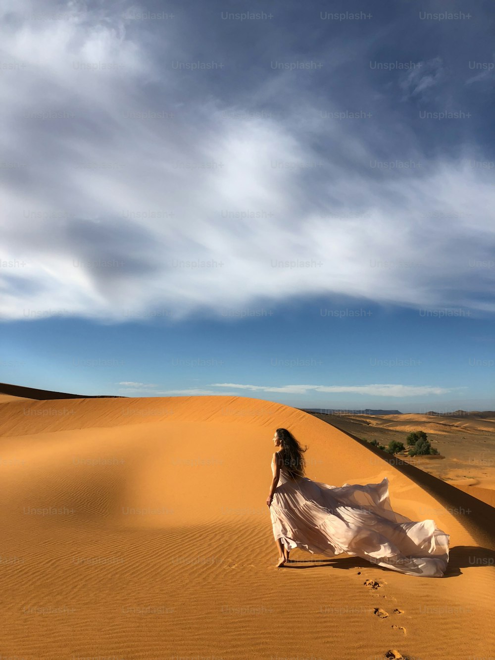 Woman in amazing silk wedding dress with fantastic view of Sahara desert sand dunes in sunset light. Landscape of Morocco, Africa.