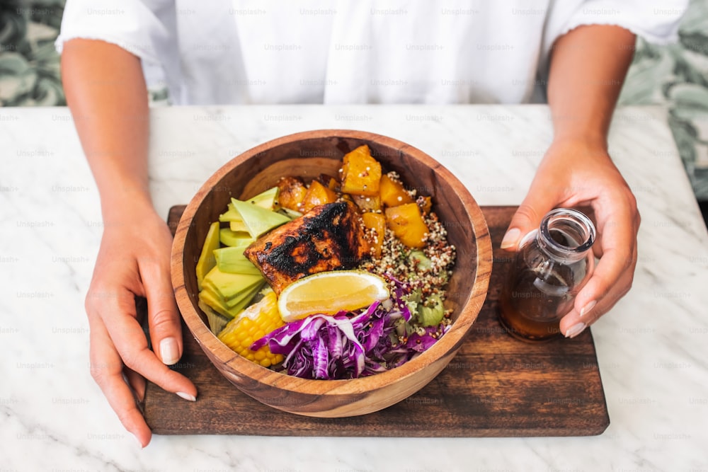 Woman eating meal with fried salmon fish steak, quinoa, avocado, corn, cabbage salad and baked pumpkin in wooden bowl. Healthy organic food concept. White marble table surface.
