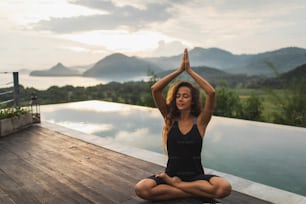 Woman meditating alone on infinity poolside with beautiful ocean and mountain view in morning. Healthy lifestyle, spiritual and emotional concept. Awakening, harmony with nature.