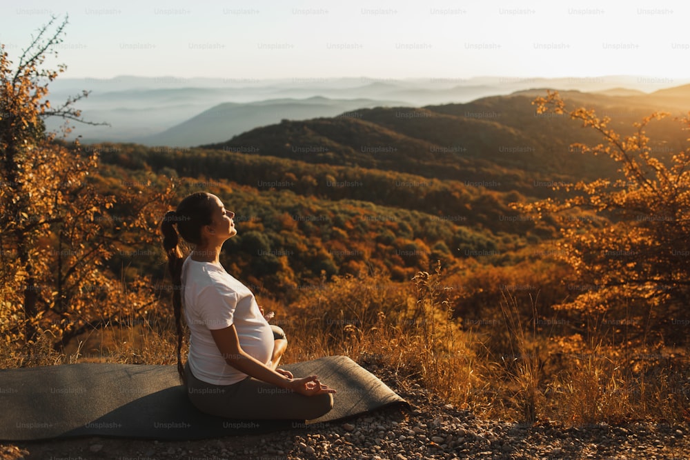 Mulher grávida em posição de lótus fazer yoga sozinha ao ar livre. Incrível vista da montanha de outono ao pôr do sol. Conceito de maternidade espiritual, harmonia natural.