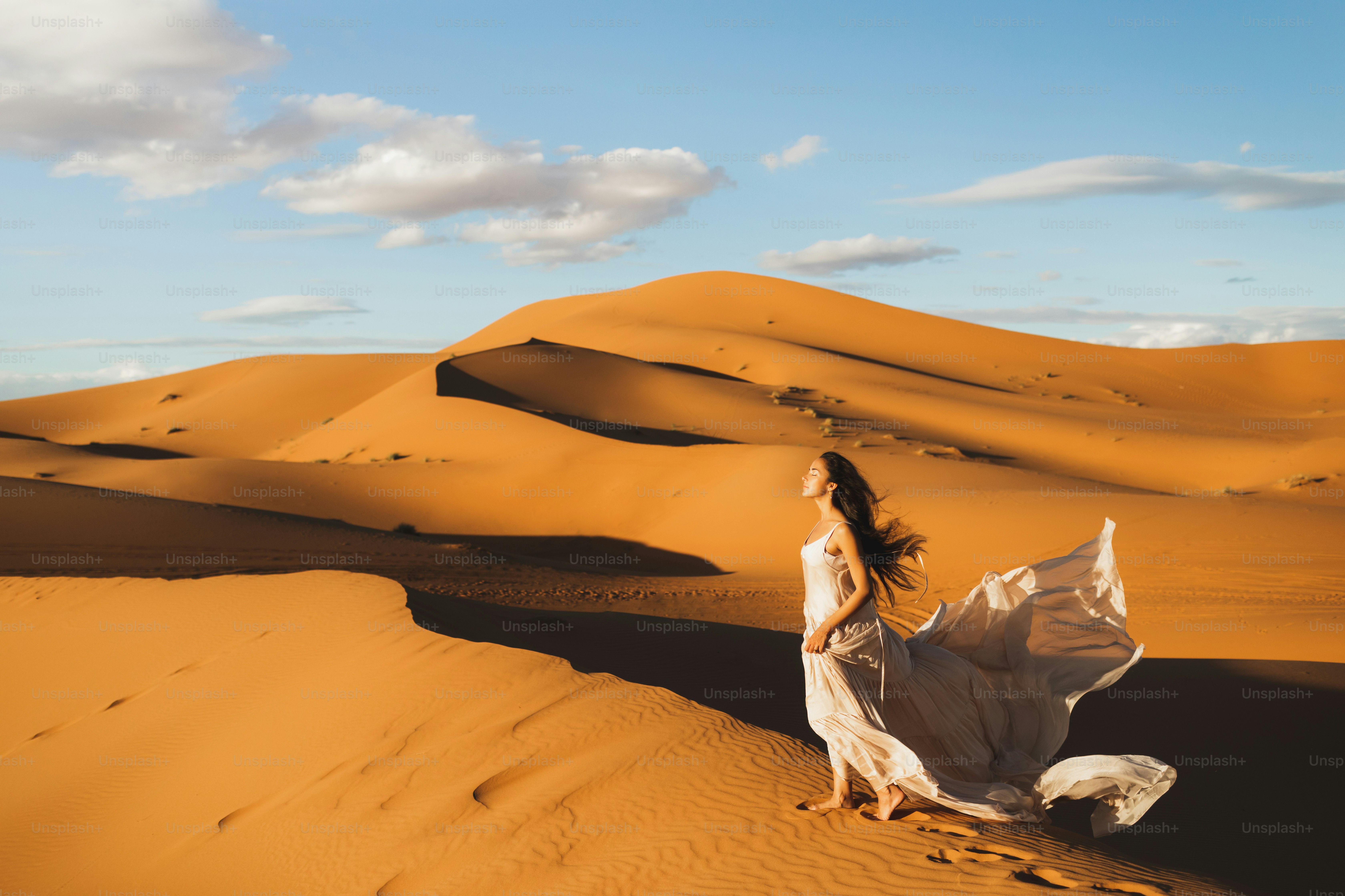 Woman in amazing silk wedding dress with fantastic view of Sahara desert sand dunes in sunset light. Landscape of Morocco, Africa.