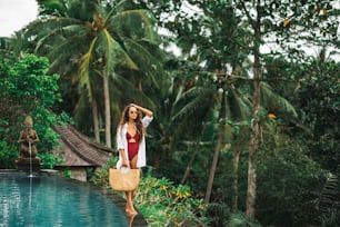 Mujer con gafas de sol, traje de baño rojo y camisa blanca relajándose y caminando junto a la piscina. Bolsa de paja hecha a mano. Piscina infinita en selva.