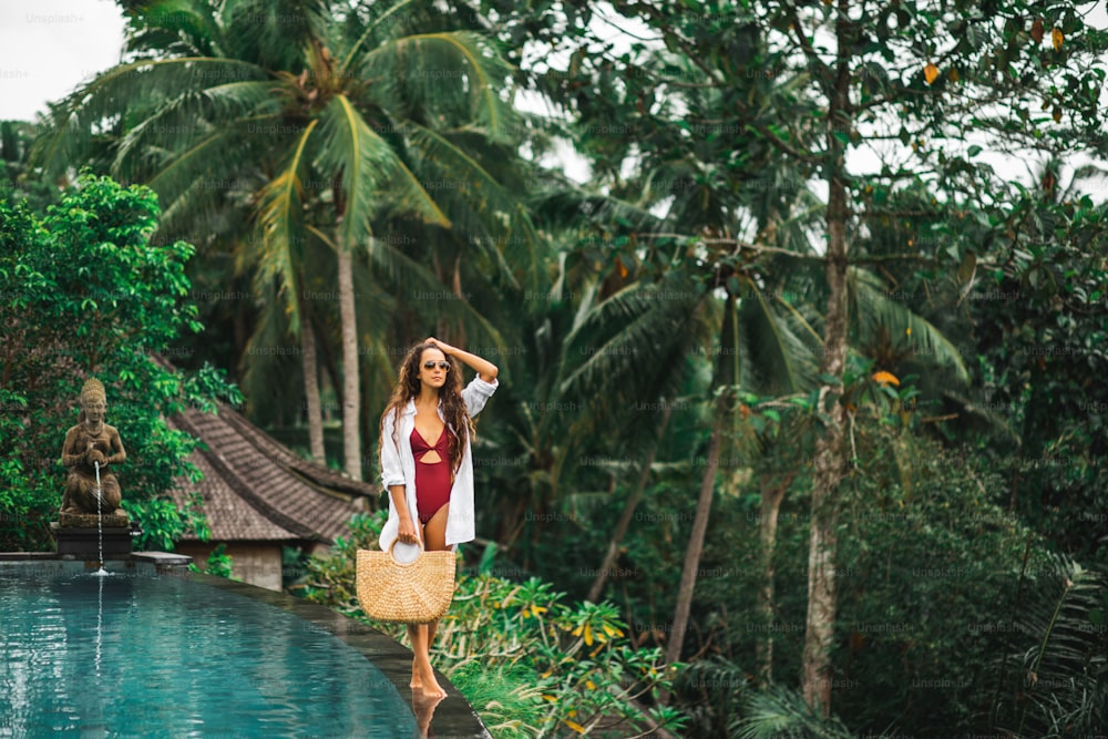 Woman in sunglasses, red swimsuit and white shirt relaxing and walking by poolside. Handmade straw bag. Infinity swimming pool in jungle.