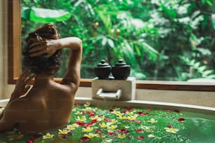 Hermosa mujer joven disfrutando en spa, vista desde atrás. Bañera de piedra de lujo con vista a la selva en ventana. Flores tropicales en el agua. Concepto de tratamiento de belleza.